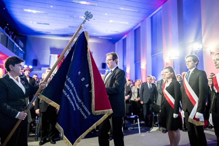 fot. P. Tracz / KPRM Premier Beata Szydło przekazuje sztandar KSAP Dyrektorowi KSAP. Z prawej strony stoi poczet sztandarowy. W tle uczestnicy uroczystości.